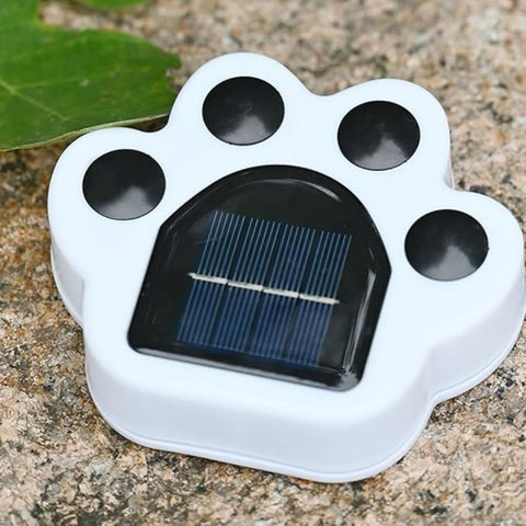 A close-up shows a small, white paw-shaped solar light sitting on a textured surface, possibly stone or concrete. The paw has black circles for the pads and toes, and a rectangular solar panel with visible grid lines is embedded in the center of the paw. A green leaf is partially visible in the upper left corner.