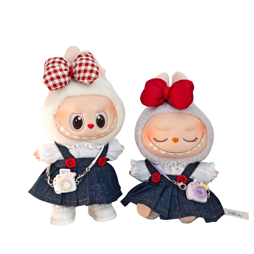 Two Labubu dolls standing side by side, both in matching denim jumper dresses with red buttons and white blouses. One wears a red bow and carries a purple camera purse, while the other has a red checkered bow and a white camera bag. Their coordinated outfits create a charming best-friend travel style.