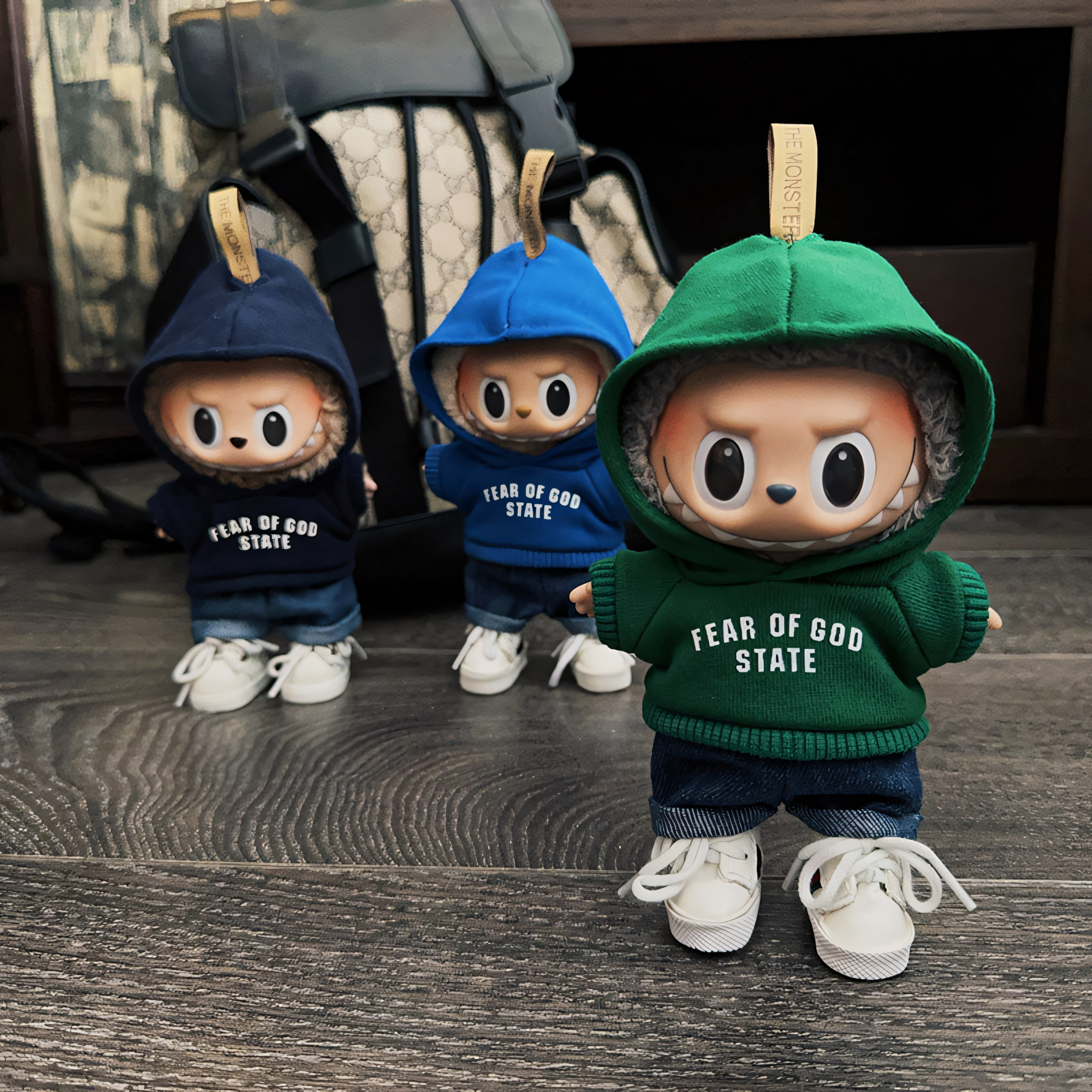 Three Labubu dolls in navy, blue, and green “FEAR OF GOD STATE” hoodies stand on a wooden floor with a luxury backpack in the background. Each doll wears jeans and white sneakers, creating a playful mini streetwear squad aesthetic.