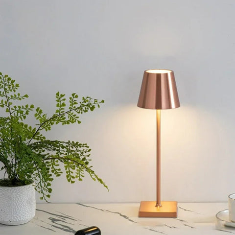 A cordless LED table lamp with a copper finish stands illuminated on a light-colored marble-patterned surface. The lamp has a tapered shade and a square base. To the left, a small white pot contains an artificial maidenhair fern with delicate green fronds. Part of a white cup and saucer are visible on the right. The background is a plain white wall.