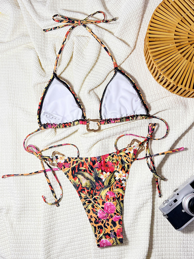 Flat lay of a floral print bikini with a halter neck top and high-cut bottoms, featuring decorative metal elements and side ties, displayed on a white towel with a woven bag and camera.