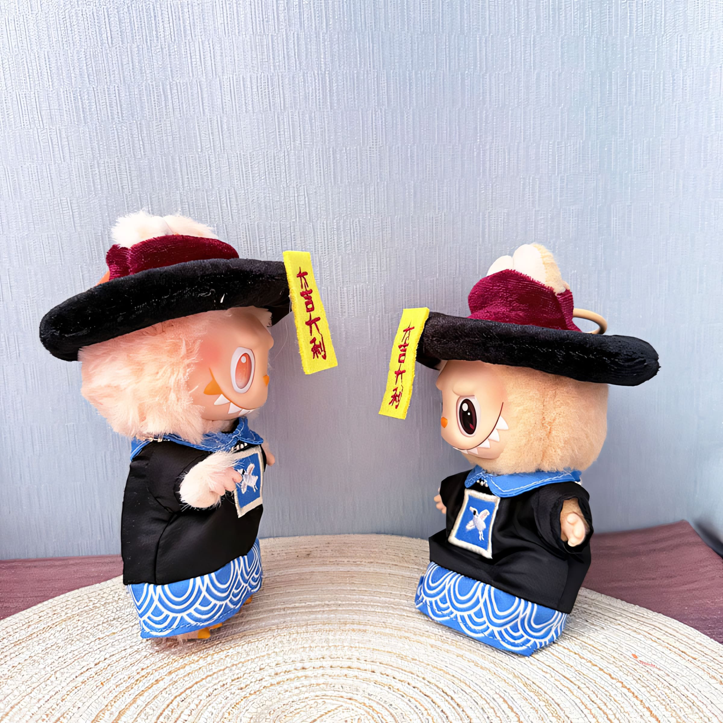 Side view of two Labubu dolls facing each other in matching Jiangshi outfits, including embroidered black robes and velvety red hats. Both dolls have yellow talismans hanging from their hats and stand on a woven circular mat with a pale blue background, capturing a fun and festive Halloween vibe.