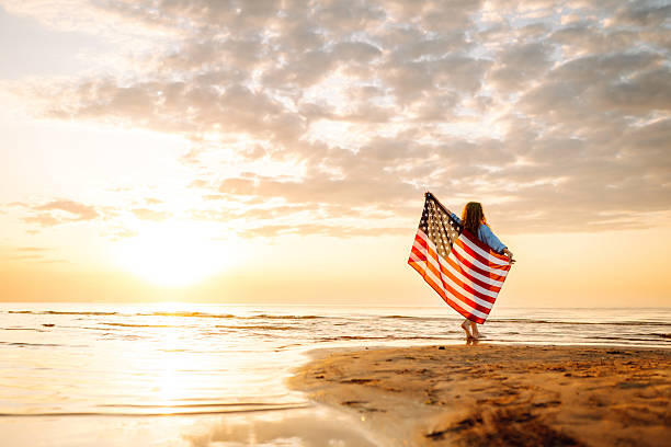 Happy woman on the beach with American flag on her shoulders. Happy woman on the beach with American flag on her shoulders. Beautiful woman enjoying sunset at sea, feeling freedom holding US flag. Independence and celebration concept. American Flag stock pictures, royalty-free photos & images
