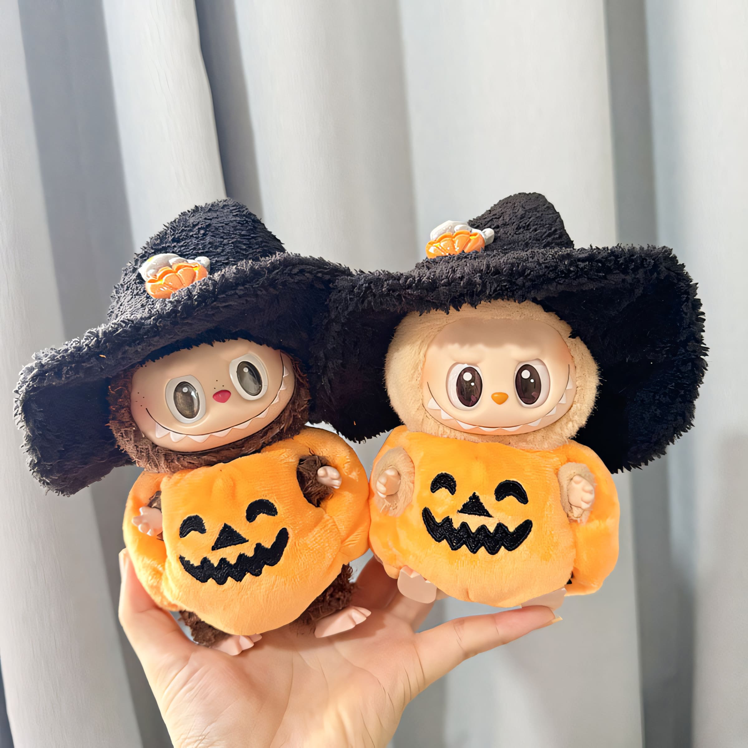Two Labubu dolls in soft jack-o'-lantern costumes with fluffy black witch hats. The matching plush pumpkin outfits are accented with embroidered faces, and the hats are topped with little pumpkin decorations, making them a charming spooky pair.