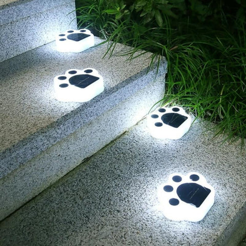 Four small, white paw-shaped solar lights are placed along the edges of outdoor steps, emitting a bright white glow from the paw pads and toes at night. The top of each light features a dark solar panel. Greenery grows along the side of the steps.