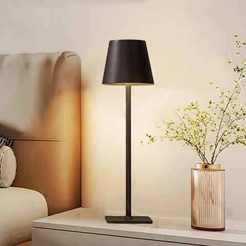 A cordless LED table lamp with a matte black tapered shade and base stands illuminated on a white nightstand. To the right, a fluted glass vase with a gold rim holds delicate artificial branches with small white flowers. Part of a light beige headboard and pillows are visible in the blurred background to the left. The wall behind is a soft beige.