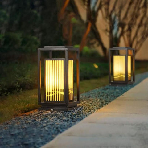 Two modern, dark-framed outdoor lanterns with vertical slats emit a warm glow along a pathway at dusk or dawn. The lanterns sit on a bed of small stones beside the paved walkway, with blurred greenery in the background.