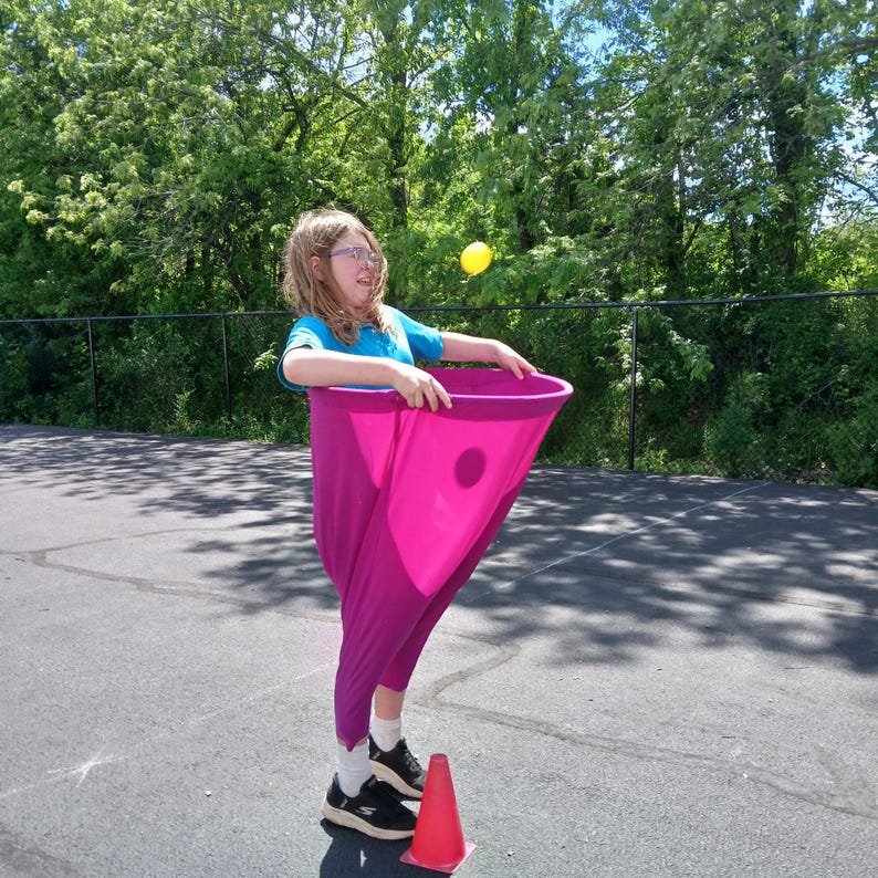 May include: A person holding a pink fabric cone with a hole in the center. A yellow ball is in the air above the cone.