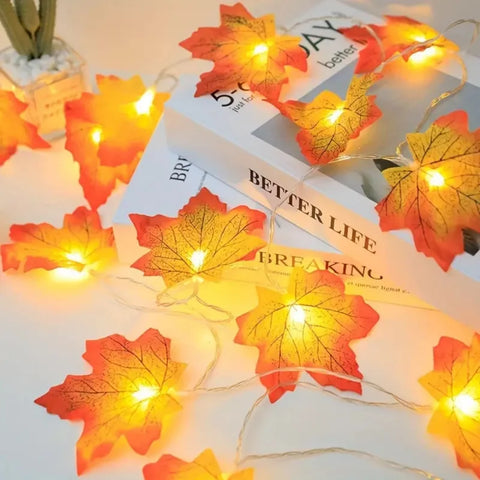 A string of warm white fairy lights shaped like autumn maple leaves is draped across a book titled "BETTER LIFE BREAKING". The artificial leaves are orange and red with visible veins, and the lights within them emit a soft glow. Part of a small potted plant is visible in the upper left corner.