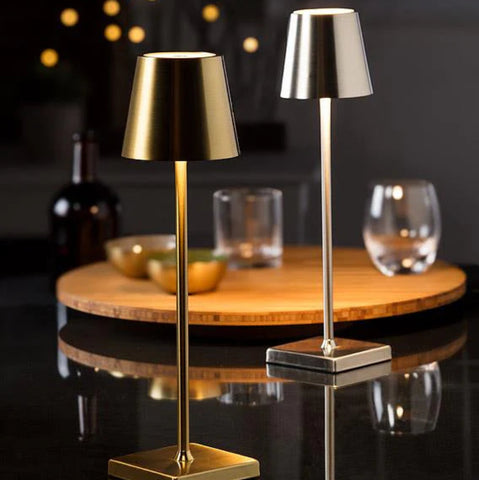 Two cordless LED table lamps with metallic finishes stand on a reflective black surface. The lamp on the left has a gold finish, while the one on the right is silver. Both have a classic lampshade shape and a square base. They emit a warm, upward light. The blurred background shows a wooden serving tray with bowls and glasses, along with a dark bottle and soft bokeh lights.