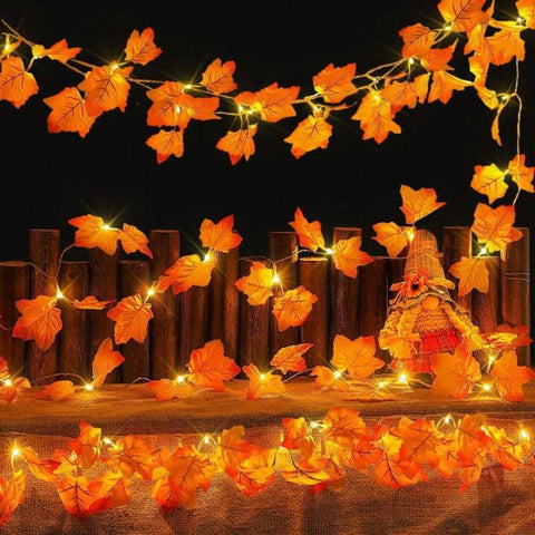 Three strands of warm white fairy lights shaped like autumn maple leaves are draped across a rustic wooden fence and a dark background. The artificial leaves are a vibrant orange, and the lights within them create a festive glow. A small, straw-like figure is positioned behind the middle strand of lights on the fence.