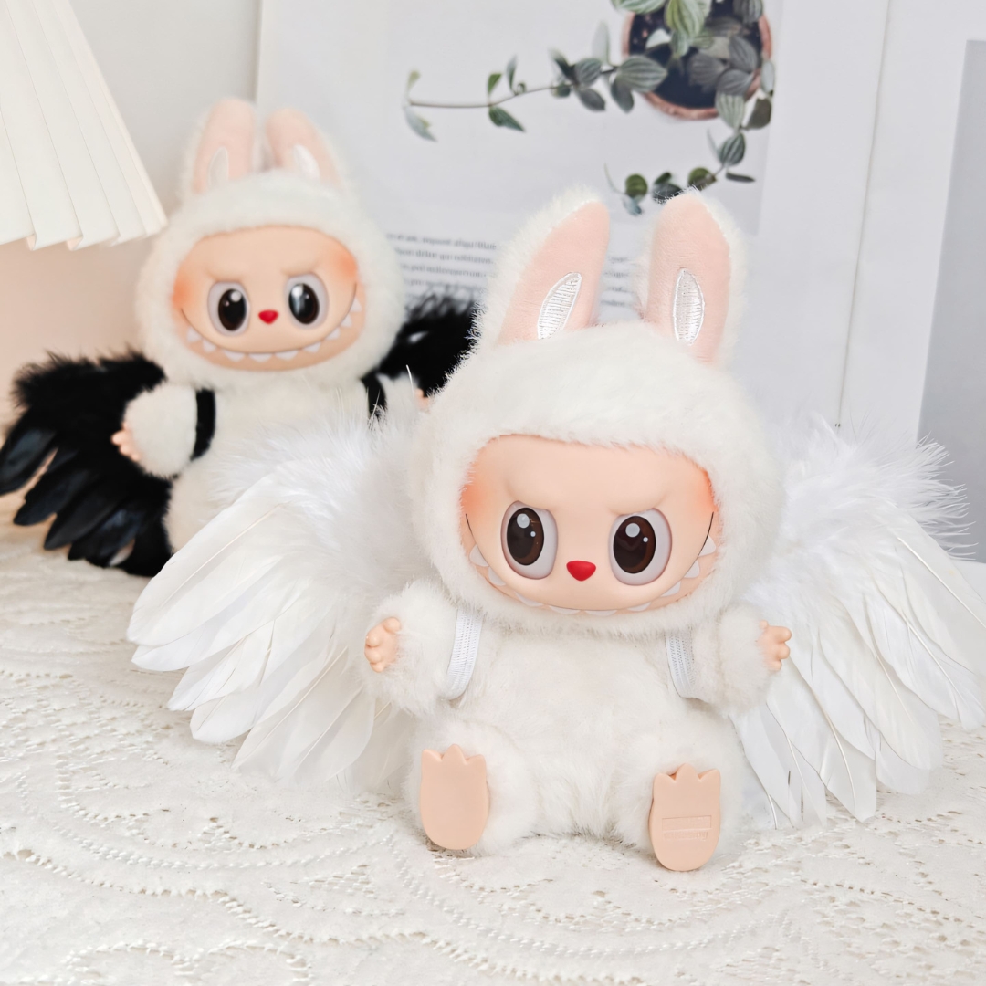 Two Labubu dolls sitting on a lace-covered surface. The one in front wears white angel wings, while the one behind features black wings. Both wear matching fluffy white hooded outfits with bunny ears, creating a playful and whimsical contrast between light and dark angel aesthetics.