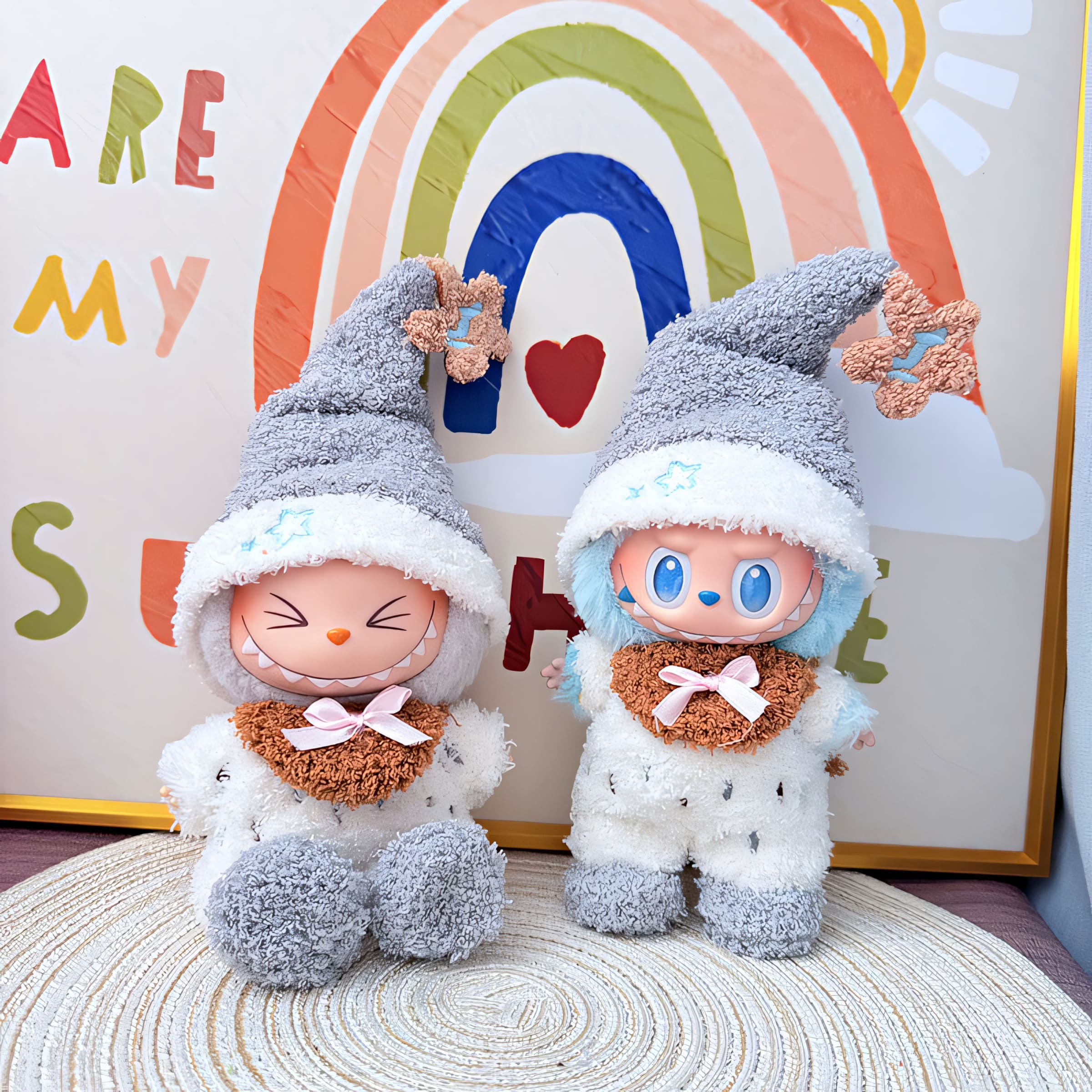 Two Labubu dolls in matching fuzzy pajama sets, featuring gray-and-white plush jumpsuits, oversized gray nightcaps with star-shaped tips, and cozy brown collars tied with pink bows. One doll has closed eyes with a warm smile, while the other has bright blue eyes and blue-tinted fur, posing cheerfully in front of a rainbow-themed "You Are My Sunshine" backdrop.